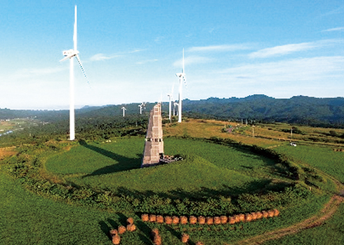 山に囲まれた牧場、中央にサイロ、周辺に複数台の風車があります。