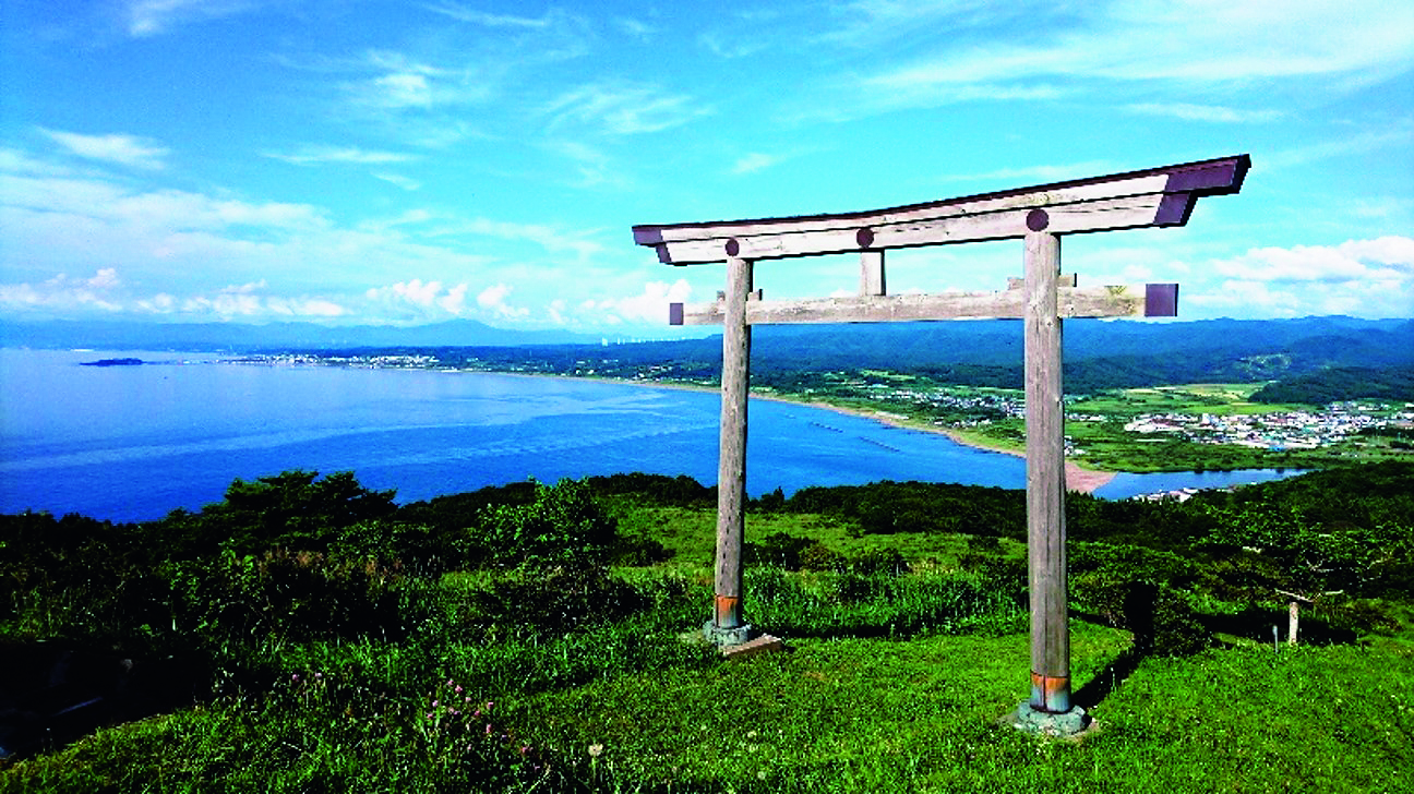 青空の下、真っ青な日本海と海岸線を一望する緑豊かな山頂。山頂には木製の鳥居があります。